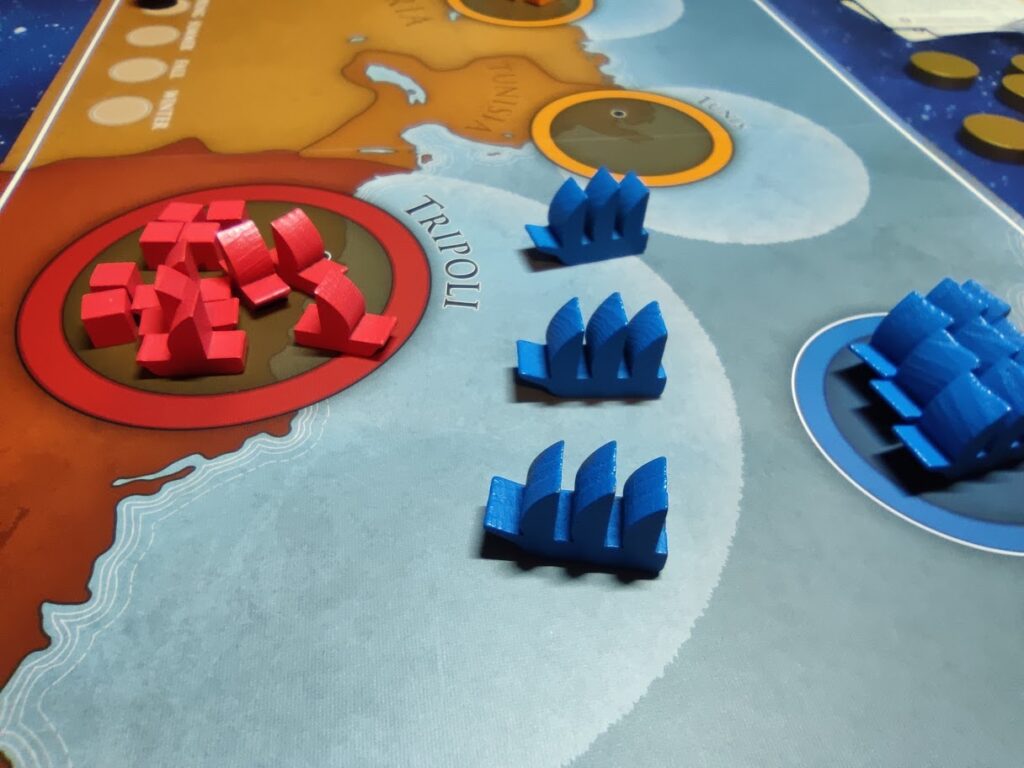 blue us frigate pieces in the centre of the image are facing the harbour of tripoli on the game board. on the left of the image are several red boats and cubes, defending tripoli