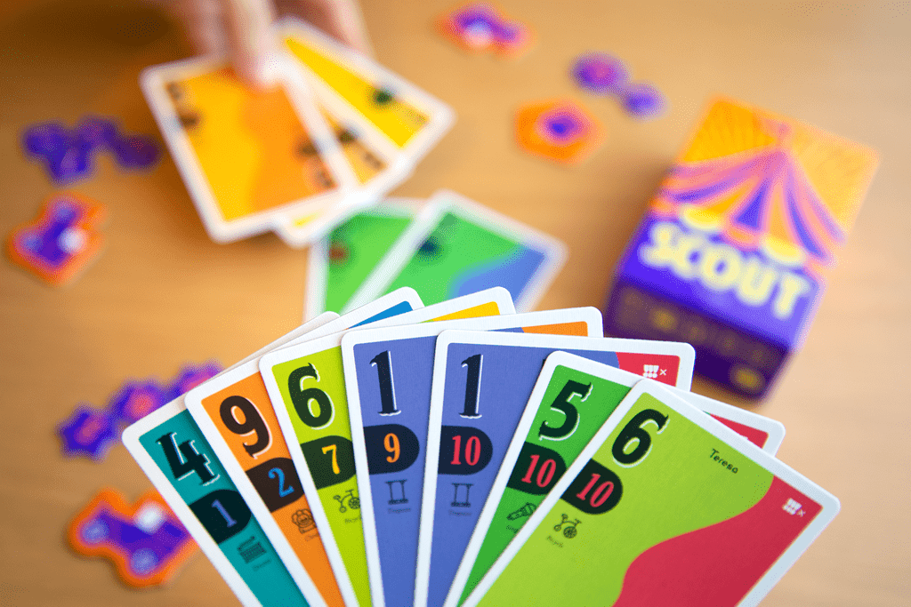 a hand of scout cards in the foreground. In the background is a blurred image of someone playing a set of cards to the table
